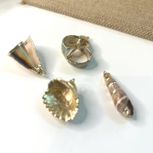 May include: An assortment of seashell-themed jewelry and decorative items. Includes a gold-toned ring, a shell-shaped pendant, a cone-shaped pendant, and a spiral shell pendant. The items are displayed on a white surface with a burlap background.