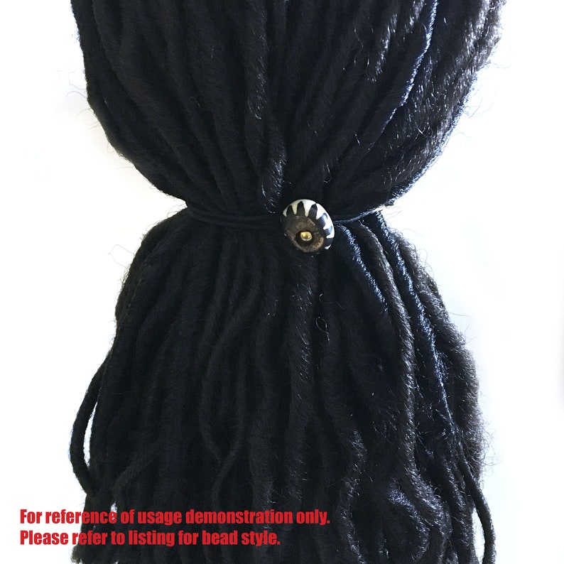 May include: A black hair tie with a white and gold bead. The bead has a geometric design.