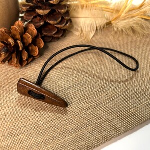May include: A wooden toggle button with a black elastic loop. The button is a dark brown color and has a smooth, polished finish. The button is designed to be used for fastening garments or other items.