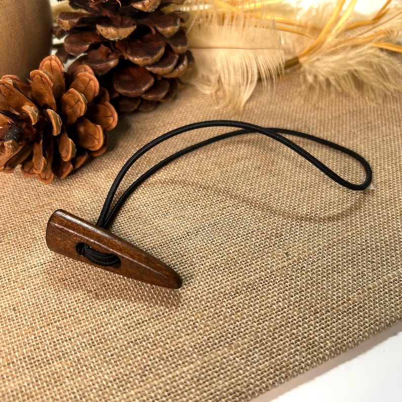 May include: A wooden toggle button with a black elastic loop. The button is a dark brown color and has a smooth, polished finish. The button is designed to be used for fastening garments or other items.