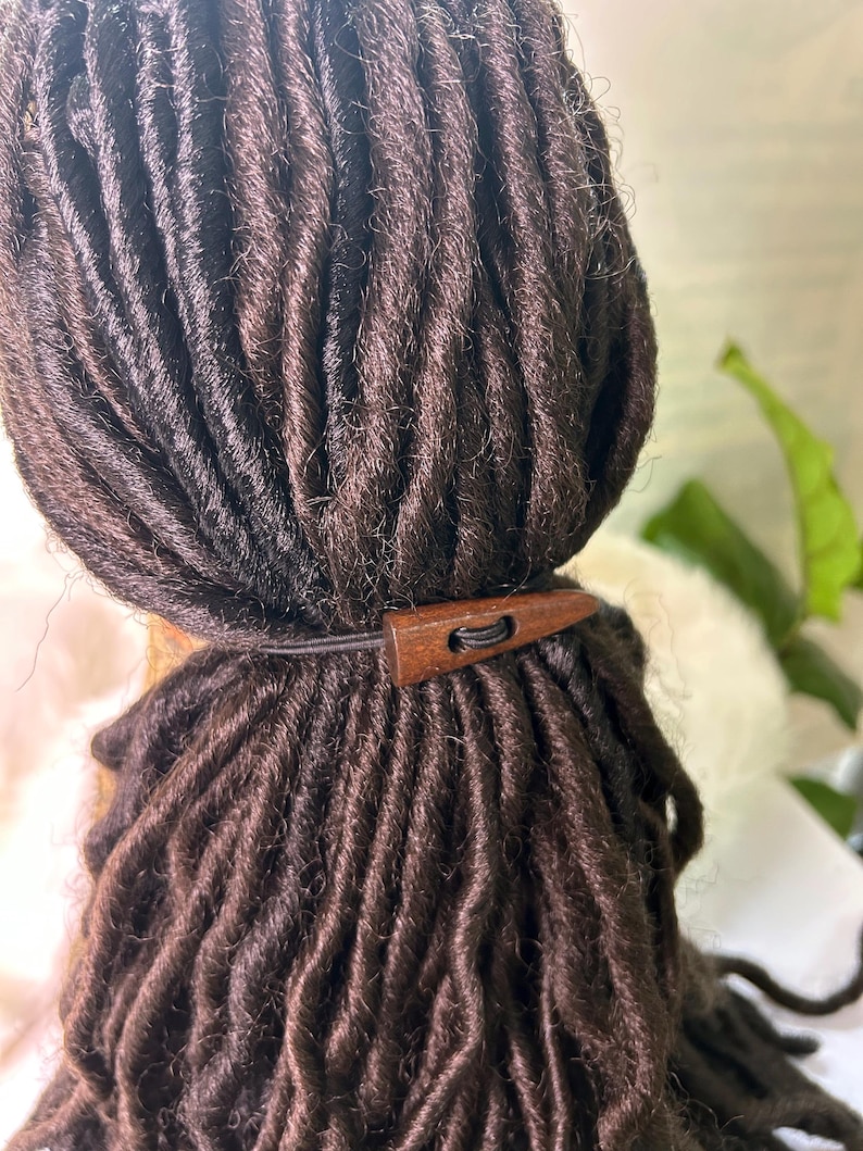 May include: Close-up of dark brown dreadlocks styled with a wooden hair accessory. The accessory is a small, rectangular piece of wood with a hole for the elastic band. The dreadlocks are neatly arranged.