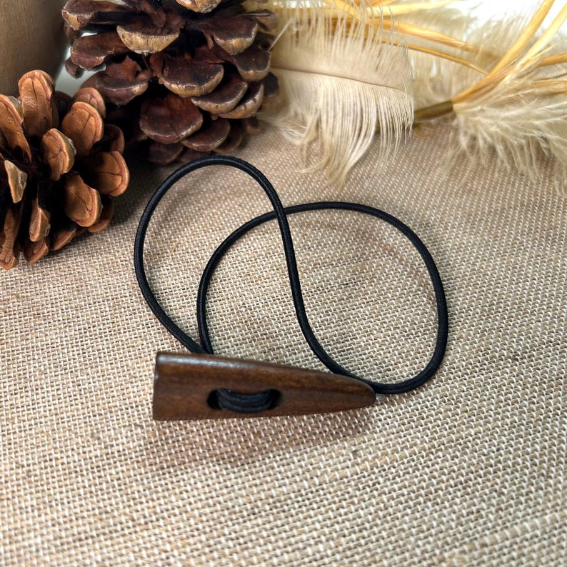 May include: A dark brown wooden hair tie with a triangular design and a black elastic band. The hair accessory is shown against a neutral background with pinecones and feathers.