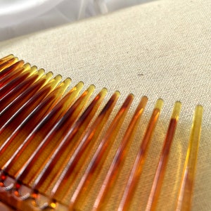May include: A close-up of a brown plastic comb with fine teeth. The comb is lying on a beige fabric surface.