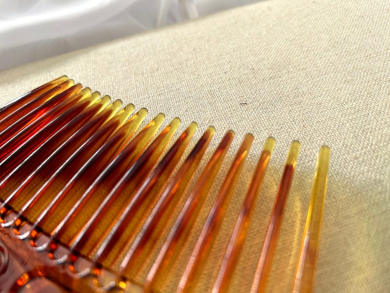 May include: A close-up of a brown plastic comb with fine teeth. The comb is lying on a beige fabric surface.