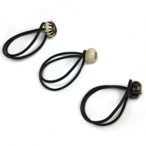 Puede incluir: Tres coleteros decorativos con bandas elásticas negras y cuentas de cerámica. Las cuentas son crema, negras y marrones, con detalles dorados. Las cuentas tienen patrones y formas únicas.