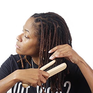 May include: A person using a light-colored wooden hair styling tool on brown dreadlocks. The tool has a black bristle brush. The person is wearing a black t-shirt. The background is white.