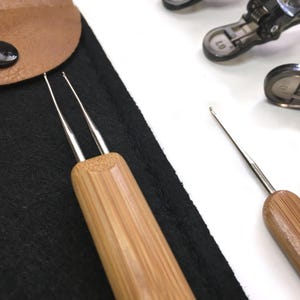 Puede incluir: Primer plano de ganchos de ganchillo con mangos de bambú. Los ganchos tienen una punta de metal plateado y se muestran cerca de una bolsa de fieltro negro. Los mangos tienen un patrón de vetas de madera natural. Otras herramientas de metal son visibles en el fondo.
