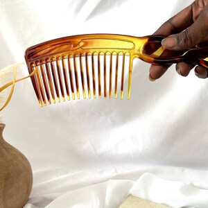 May include: A brown plastic comb with a curved handle. The comb has a wide tooth design and is held in a hand.