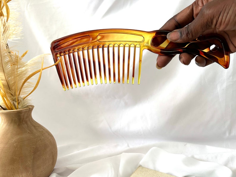 May include: A brown plastic comb with a curved handle. The comb has a wide tooth design and is held in a hand.