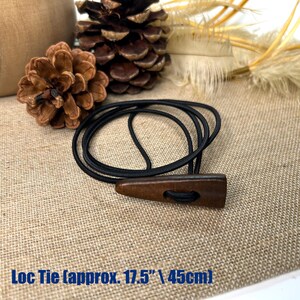 May include: A black elastic Loc Tie with a dark brown wooden toggle, approximately 17.5 inches (45cm) long. The toggle has a hole for the elastic to pass through. The background includes pinecones and dried plants.
