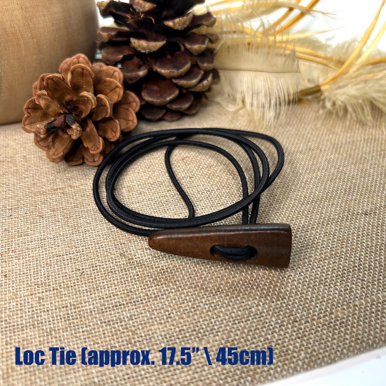 May include: A black elastic Loc Tie with a dark brown wooden toggle, approximately 17.5 inches (45cm) long. The toggle has a hole for the elastic to pass through. The background includes pinecones and dried plants.
