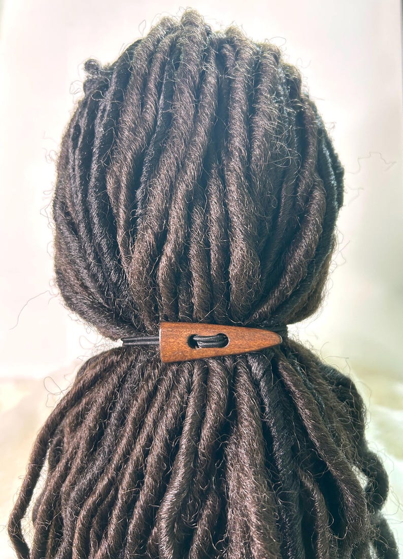 May include: Dark brown dreadlocks styled with a wooden hair accessory. The accessory is a light brown color and is secured with a black elastic band. The dreadlocks are tightly wound and have a natural texture.