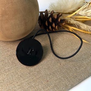 May include: A glossy black bottle stopper with a round design and a black rubber cord. The stopper is on a tan surface with a wooden sphere and dried plants in the background. The stopper is a decorative item.