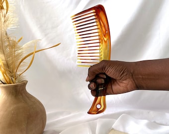 Può includere: Un pettine di plastica marrone e ambrato con denti larghi. Il pettine è tenuto in una mano.