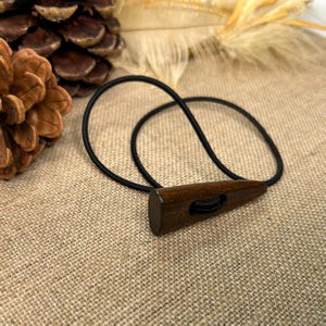 May include: A dark brown wooden toggle button with a black elastic loop. The button is triangular with a rounded end and a hole for the elastic. The background is a textured tan fabric.