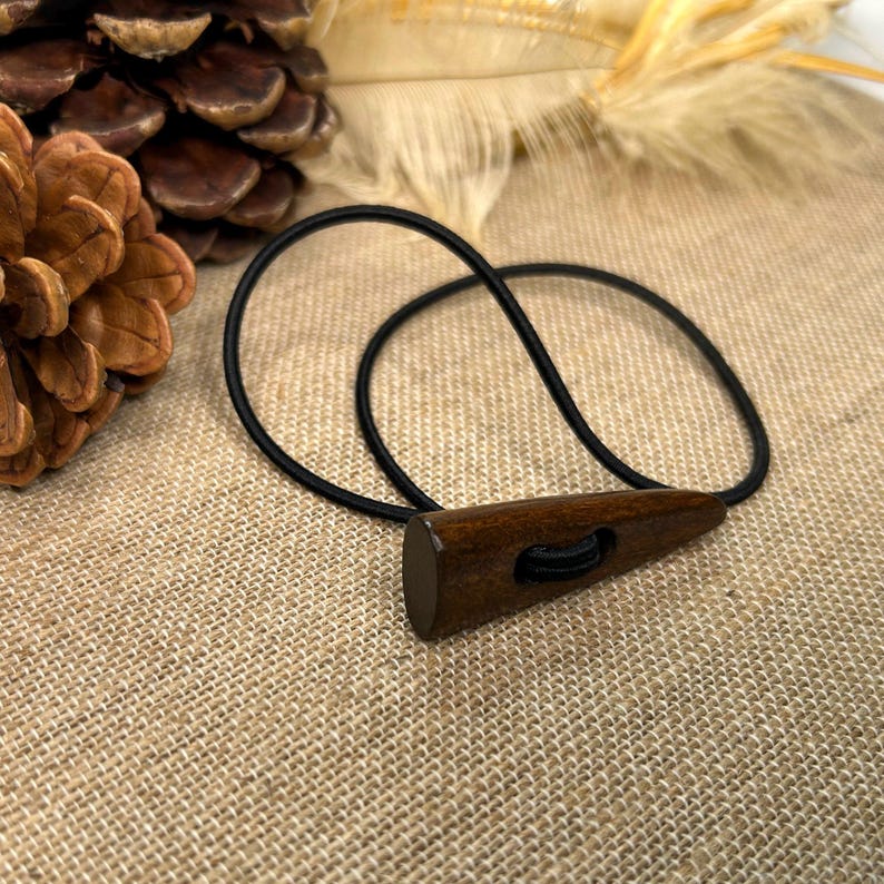 May include: A dark brown wooden toggle button with a black elastic loop. The button is triangular with a rounded end and a hole for the elastic. The background is a textured tan fabric.