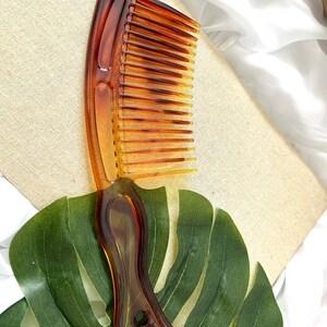 May include: A brown plastic comb with a wide tooth design and a curved handle. The comb is lying on a green leaf.