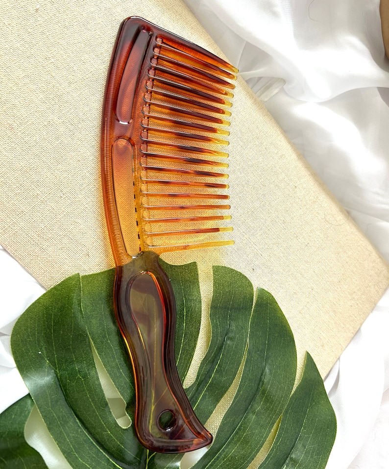May include: A brown plastic comb with a wide tooth design and a curved handle. The comb is lying on a green leaf.