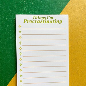 May include: A white notepad with the text "Things I'm Procrastinating" in green. The notepad has lines for writing and is decorated with gold starbursts. The background is a split of green and gold.