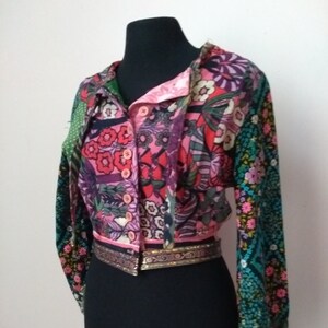 Può includere: Una blusa con stampa floreale multicolore con uno schema di colori rosa, verde e nero. La blusa ha un davanti abbottonato, maniche lunghe e un bordo dorato intorno alla vita.