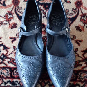 Puede incluir: Un par de zapatos de cuero negro con punta puntiaguda y una correa sobre el empeine. Los zapatos tienen un patr&oacute;n decorativo de peque&ntilde;os agujeros y un nombre de marca en el interior.