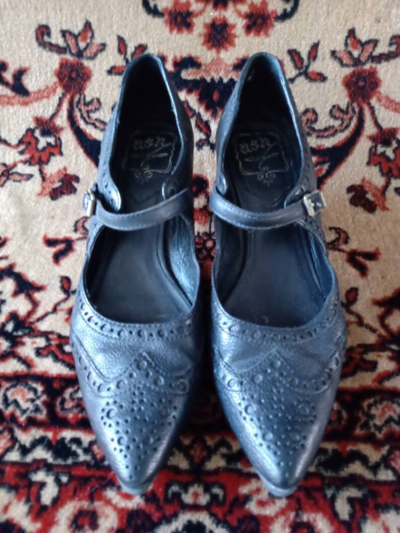 Puede incluir: Un par de zapatos de cuero negro con punta puntiaguda y una correa sobre el empeine. Los zapatos tienen un patr&oacute;n decorativo de peque&ntilde;os agujeros y un nombre de marca en el interior.