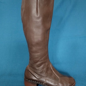 Puede incluir: Una bota alta de cuero marrón, con punta redonda y tacón bajo. La bota presenta una suela cosida y una parte superior de cuero lisa, ligeramente arrugada. La bota está diseñada para llegar a la rodilla y tiene un diseño sencillo y clásico.
