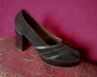 May include: Black platform heels with a thick block heel. The shoes have a rounded toe and a single strap across the top.