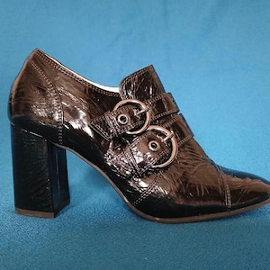Puede incluir: Botín negro de charol con tacón ancho. El zapato presenta dos cierres de hebilla plateados y una puntera en punta. El tacón mide aproximadamente 7,6 cm de alto.