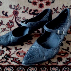 Puede incluir: Un par de zapatos de cuero negro con punta puntiaguda y una correa de hebilla. Los zapatos tienen un dise&ntilde;o de brogue y est&aacute;n sobre una alfombra estampada.