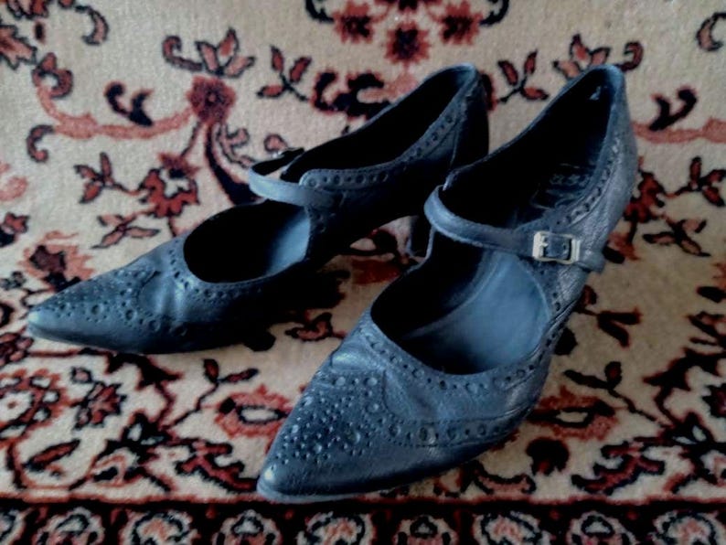 Puede incluir: Un par de zapatos de cuero negro con punta puntiaguda y una correa de hebilla. Los zapatos tienen un dise&ntilde;o de brogue y est&aacute;n sobre una alfombra estampada.