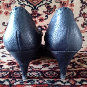 Puede incluir: Un par de zapatos de tac&oacute;n de cuero negro con punta puntiaguda y un detalle de costura decorativa en la parte posterior.
