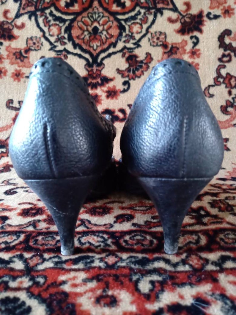 Puede incluir: Un par de zapatos de tac&oacute;n de cuero negro con punta puntiaguda y un detalle de costura decorativa en la parte posterior.