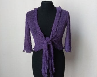 May include: A purple knitted cardigan with a tie front and ruffled edges. The cardigan is on a black mannequin.