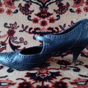 Puede incluir: Un zapato de tac&oacute;n de cuero negro con punta puntiaguda y un patr&oacute;n de brogue decorativo. El zapato tiene una correa sobre el empeine y un tac&oacute;n peque&ntilde;o.