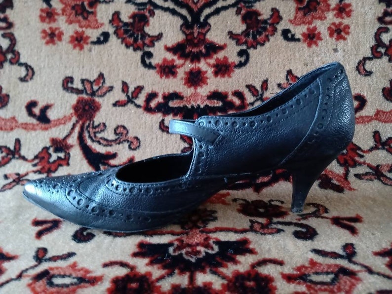Puede incluir: Un zapato de tac&oacute;n de cuero negro con punta puntiaguda y un patr&oacute;n de brogue decorativo. El zapato tiene una correa sobre el empeine y un tac&oacute;n peque&ntilde;o.