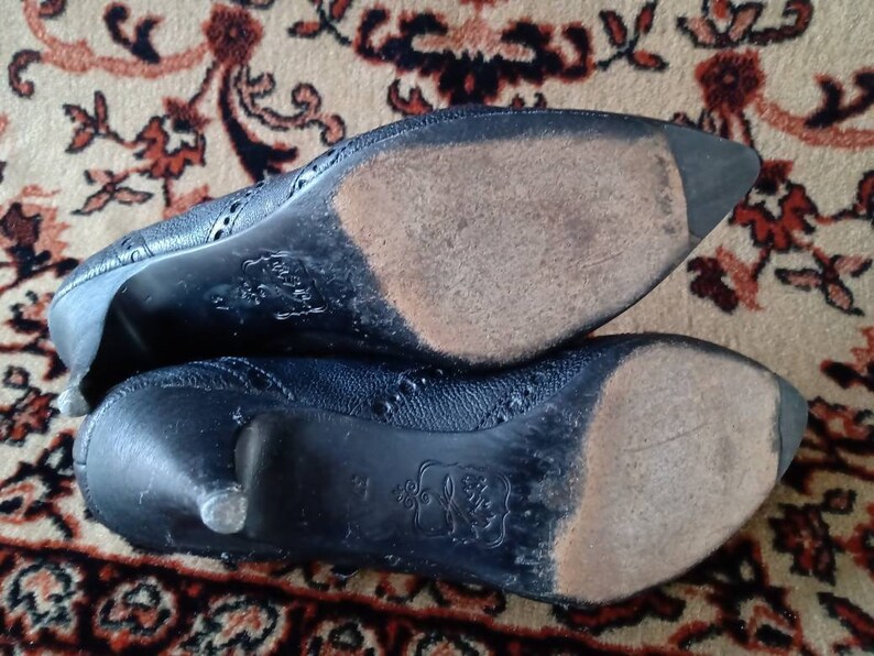 Puede incluir: Primer plano de las suelas de dos zapatos de tac&oacute;n de cuero negro. Los zapatos tienen un patr&oacute;n decorativo y un aspecto desgastado.