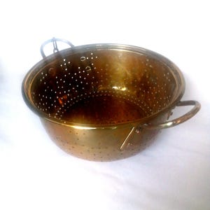Puede incluir: Un colador de latón vintage con dos asas y un fondo perforado. El colador es un utensilio de cocina que se utiliza para escurrir los alimentos.