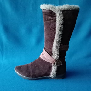 Könnte beinhalten: Braune Wildlederstiefel mit einem Kunstfellbesatz am oberen Rand des Stiefels. Die Stiefel haben einen rosafarbenen Riemen und ein Schnallendetail.
