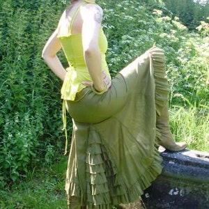 May include: A woman wearing a green skirt with ruffles and a yellow tank top. She is standing on a stone wall with green grass and plants in the background.