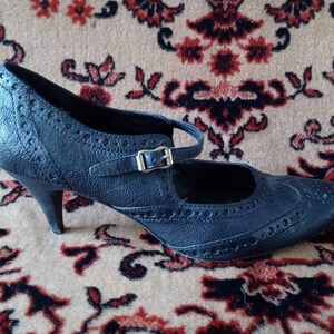 Puede incluir: Zapato de tac&oacute;n de cuero negro con punta puntiaguda y correa con hebilla. El zapato tiene un dise&ntilde;o de brogue.