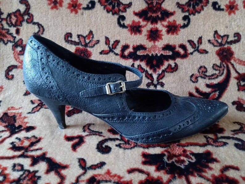 Puede incluir: Zapato de tac&oacute;n de cuero negro con punta puntiaguda y correa con hebilla. El zapato tiene un dise&ntilde;o de brogue.