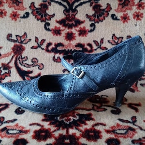 Puede incluir: Zapato de tac&oacute;n de cuero negro con punta puntiaguda y una correa con hebilla. El zapato tiene un dise&ntilde;o de brogue.