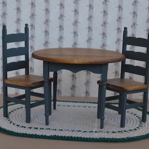 May include: A miniature dollhouse dining set with a round wooden table and two chairs. The table and chairs are painted a dark blue-green color with wooden seats. The set is placed on a white and green oval rug.