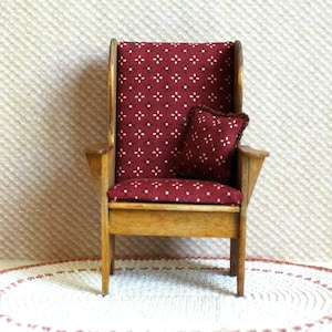 May include: Miniature wooden armchair with a burgundy seat and backrest, patterned with small white crosses. A matching pillow rests on the seat. The chair has a light brown wooden frame and armrests, and is a dollhouse furniture item.
