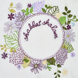 May include: Embroidered white fabric with a wreath of purple, green, and yellow flowers surrounding a circle with the text "Shabbat Shalom" in purple thread.