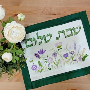 Peut inclure: Un chemin de table brodé blanc avec une bordure verte. Le chemin de table présente un motif floral avec des fleurs violettes et vertes et le texte hébreu "Shabbat Shalom".