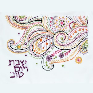 May include: White fabric with colorful paisley embroidery. The embroidery features swirls, dots, and zigzags in shades of pink, orange, green, and purple. The Hebrew text "Shabbat Shalom" is embroidered in purple in the lower left corner.