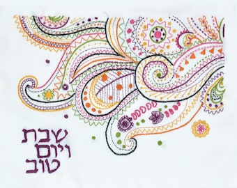 May include: White fabric with colorful paisley embroidery. The embroidery features swirls, dots, and zigzags in shades of pink, orange, green, and purple. The Hebrew text "Shabbat Shalom" is embroidered in purple in the lower left corner.