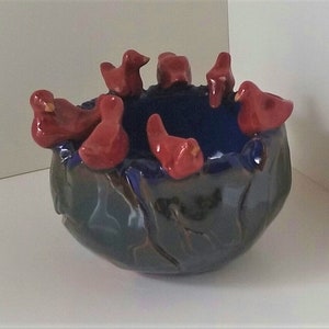 May include: A ceramic bowl with a dark blue interior and exterior, adorned with several red bird figurines around the rim. The bowl has a glossy finish and sits on a white surface.